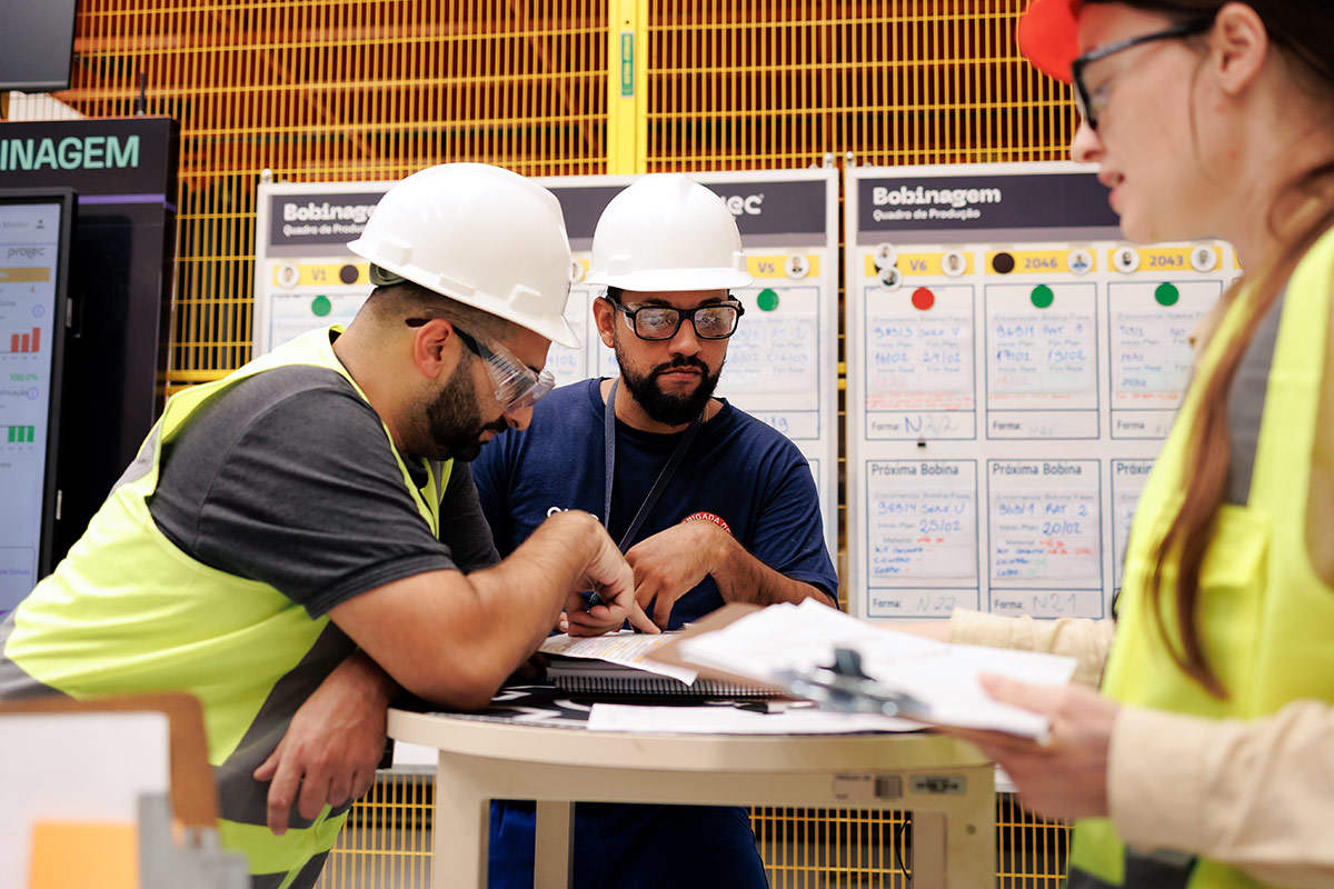 GE Vernova Kaizen Week, three workers wearing hard hats, safety vests and safety glasses stand at a table reviewing documents.
