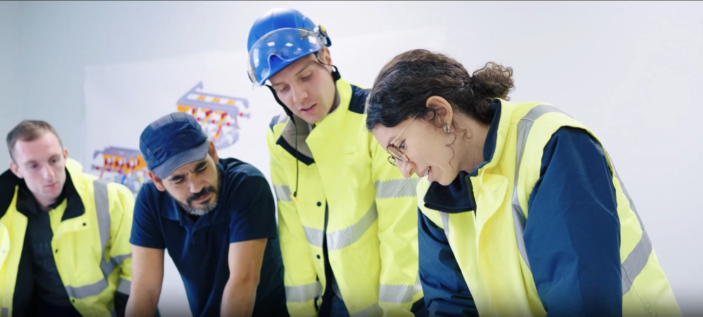 Four people in yellow work vests and jackets leaning over a table working