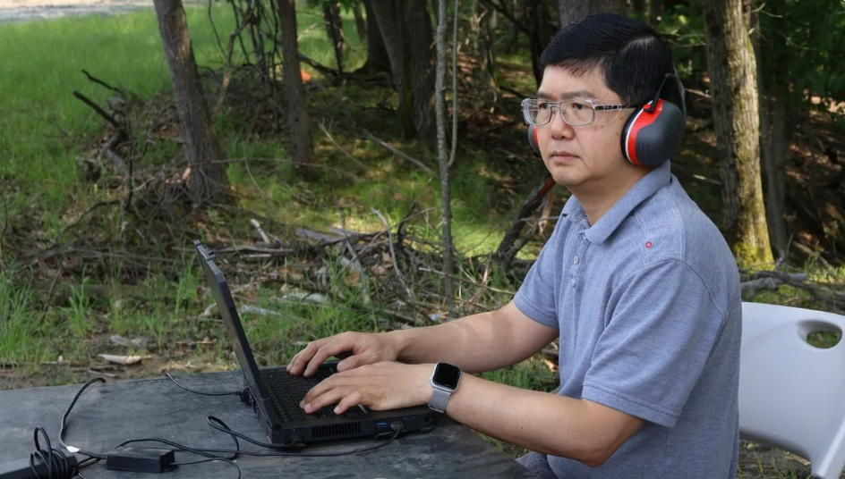 Asian man in blue polo, glasses, and headphone sitting at a computer outdoors